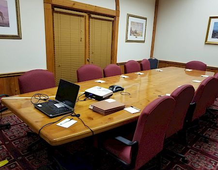 A conference room with a long wooden table, laptops and papers, purple chairs, blinds, and framed landscape paintings on the walls.