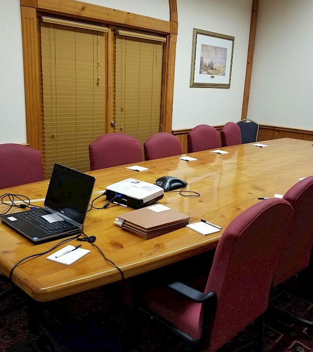 A conference room with a long wooden table, laptops and papers, purple chairs, blinds, and framed landscape paintings on the walls.