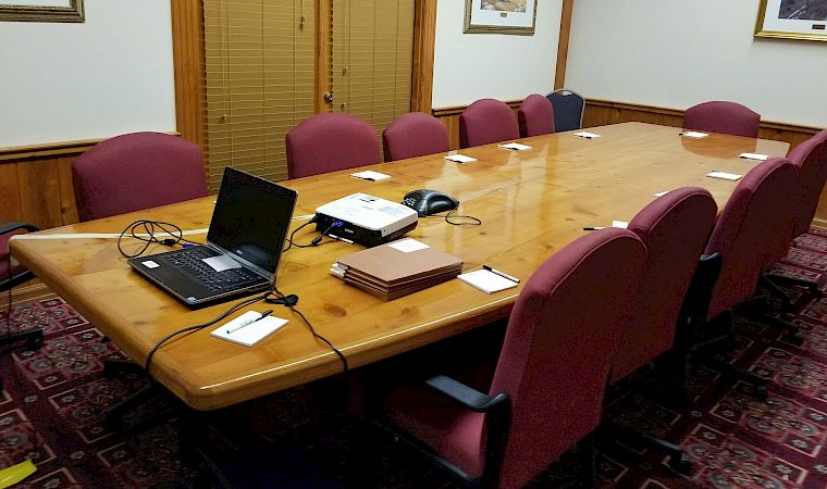 A conference room with a long wooden table, laptops and papers, purple chairs, blinds, and framed landscape paintings on the walls.