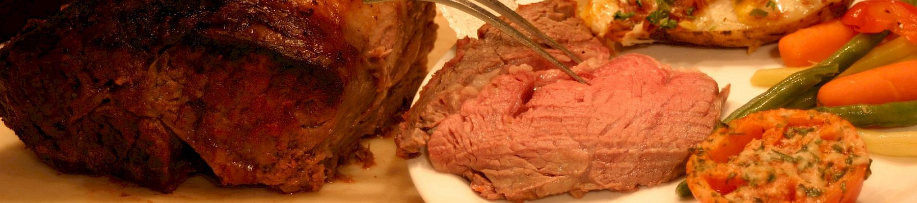 A chef carves grilled meat on a platter while a plate of steak, roasted vegetables, and herb-topped sides sits ready.