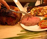 A chef carves grilled meat on a platter while a plate of steak, roasted vegetables, and herb-topped sides sits ready.