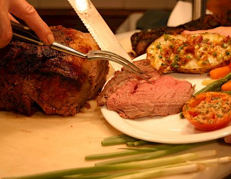 A chef carves grilled meat on a platter while a plate of steak, roasted vegetables, and herb-topped sides sits ready.