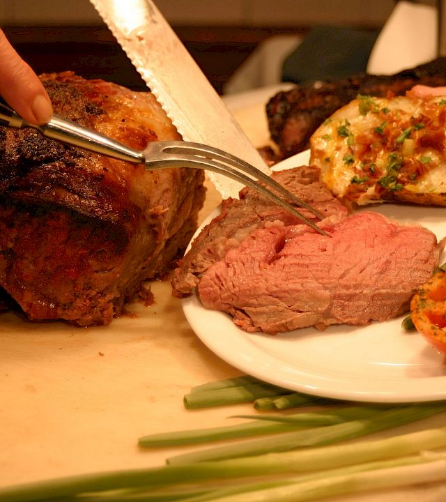 A chef carves grilled meat on a platter while a plate of steak, roasted vegetables, and herb-topped sides sits ready.