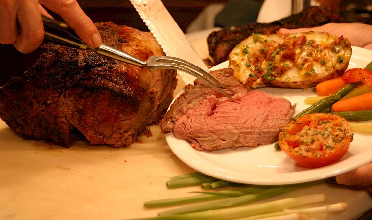 A chef carves grilled meat on a platter while a plate of steak, roasted vegetables, and herb-topped sides sits ready.
