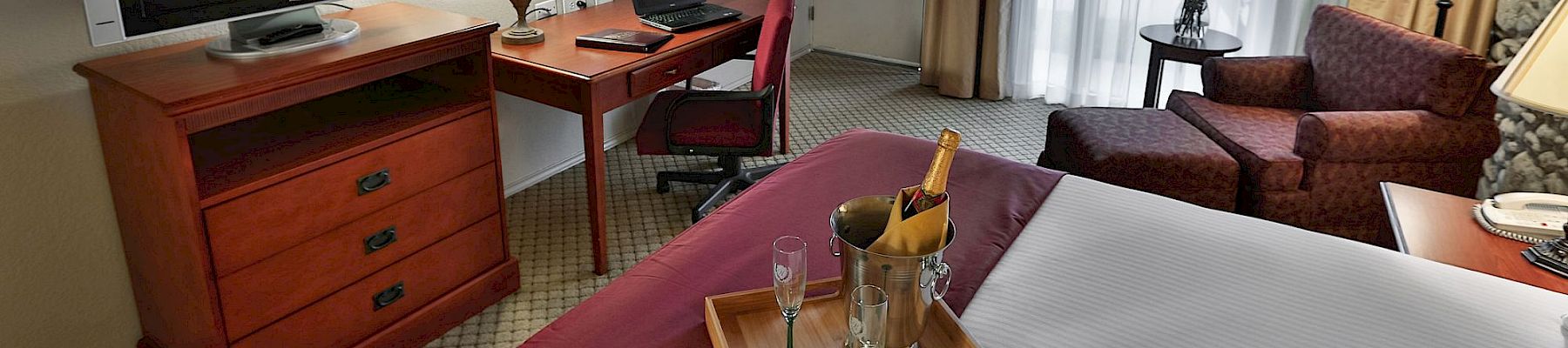 A hotel room office area with a desk, TV, laptop, lamps, curtains, sofa, and a small table with a champagne bucket.