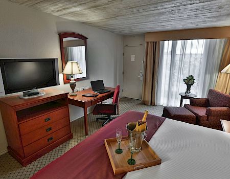 A hotel room office area with a desk, TV, laptop, lamps, curtains, sofa, and a small table with a champagne bucket.