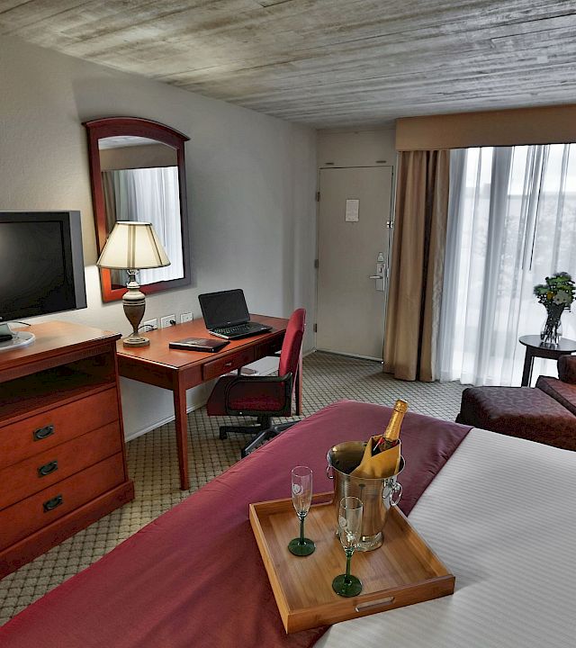 A hotel room office area with a desk, TV, laptop, lamps, curtains, sofa, and a small table with a champagne bucket.