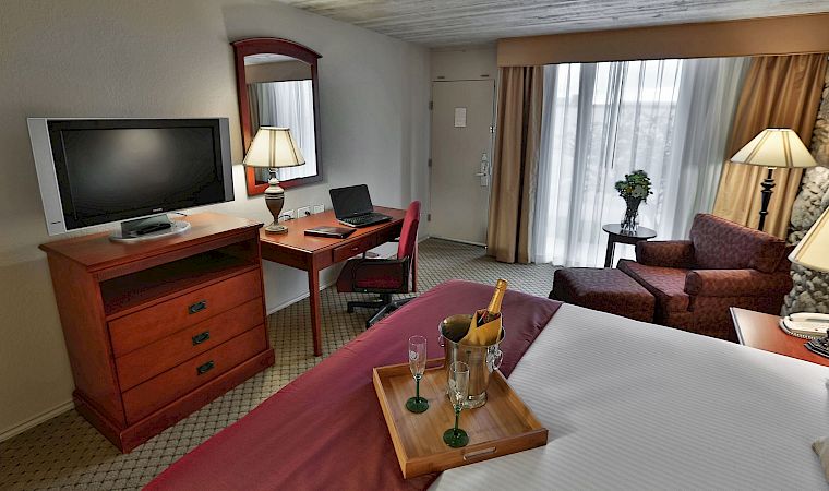 A hotel room office area with a desk, TV, laptop, lamps, curtains, sofa, and a small table with a champagne bucket.
