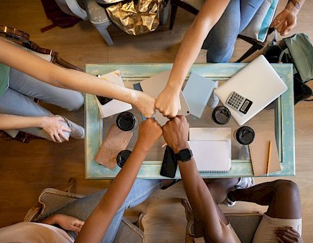 People sit around a table collaborating with laptops, notebooks, and coffee while planning or working together in a casual meeting.