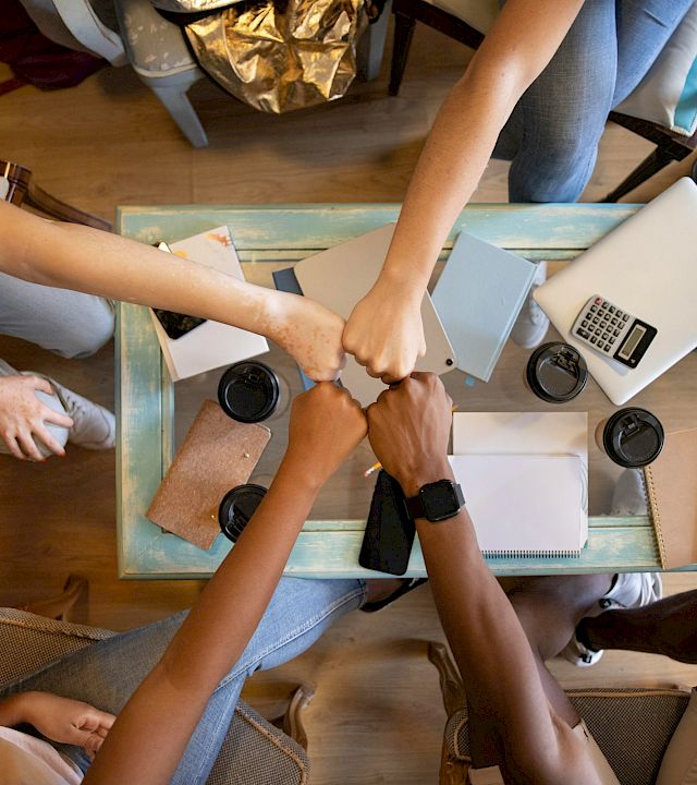 People sit around a table collaborating with laptops, notebooks, and coffee while planning or working together in a casual meeting.