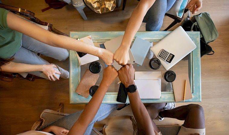 People sit around a table collaborating with laptops, notebooks, and coffee while planning or working together in a casual meeting.