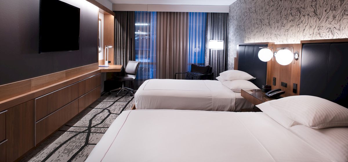 A modern hotel room with two neatly made beds, warm lighting, and dark wood accents with curtains at the window.