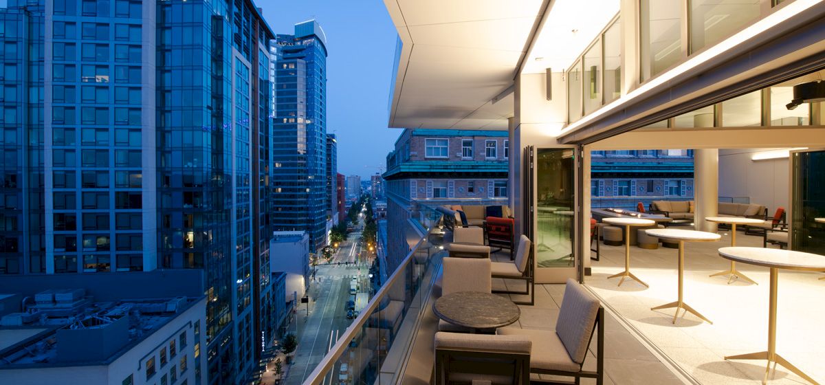 A rooftop terrace with lounge seating overlooks a modern city skyline and streets at dusk.