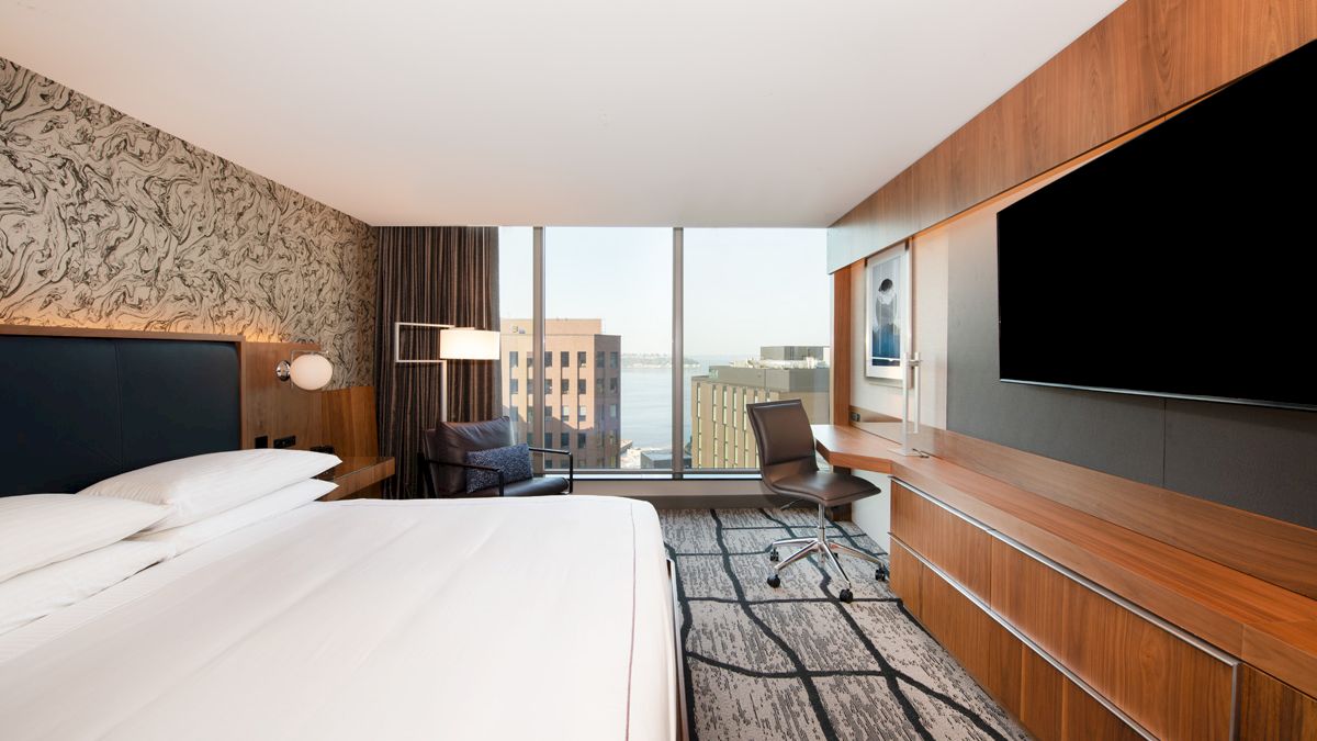 A modern hotel room with a large bed, desk chair, wall TV, sleek wood cabinetry, and big windows overlooking city buildings.