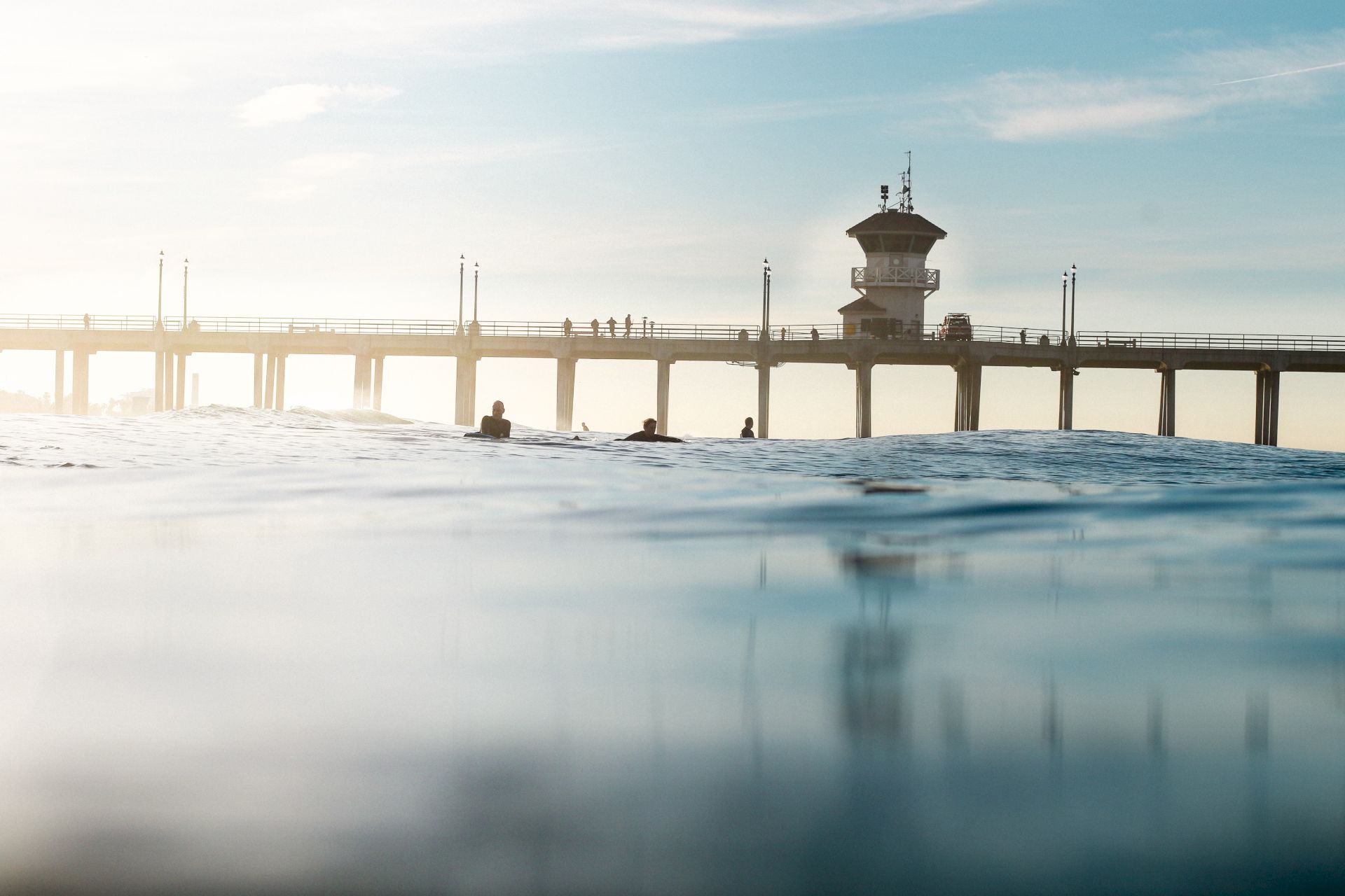 People swim in choppy water beside a long pier, with a lifeguard tower and poles silhouetted against a pale sky.